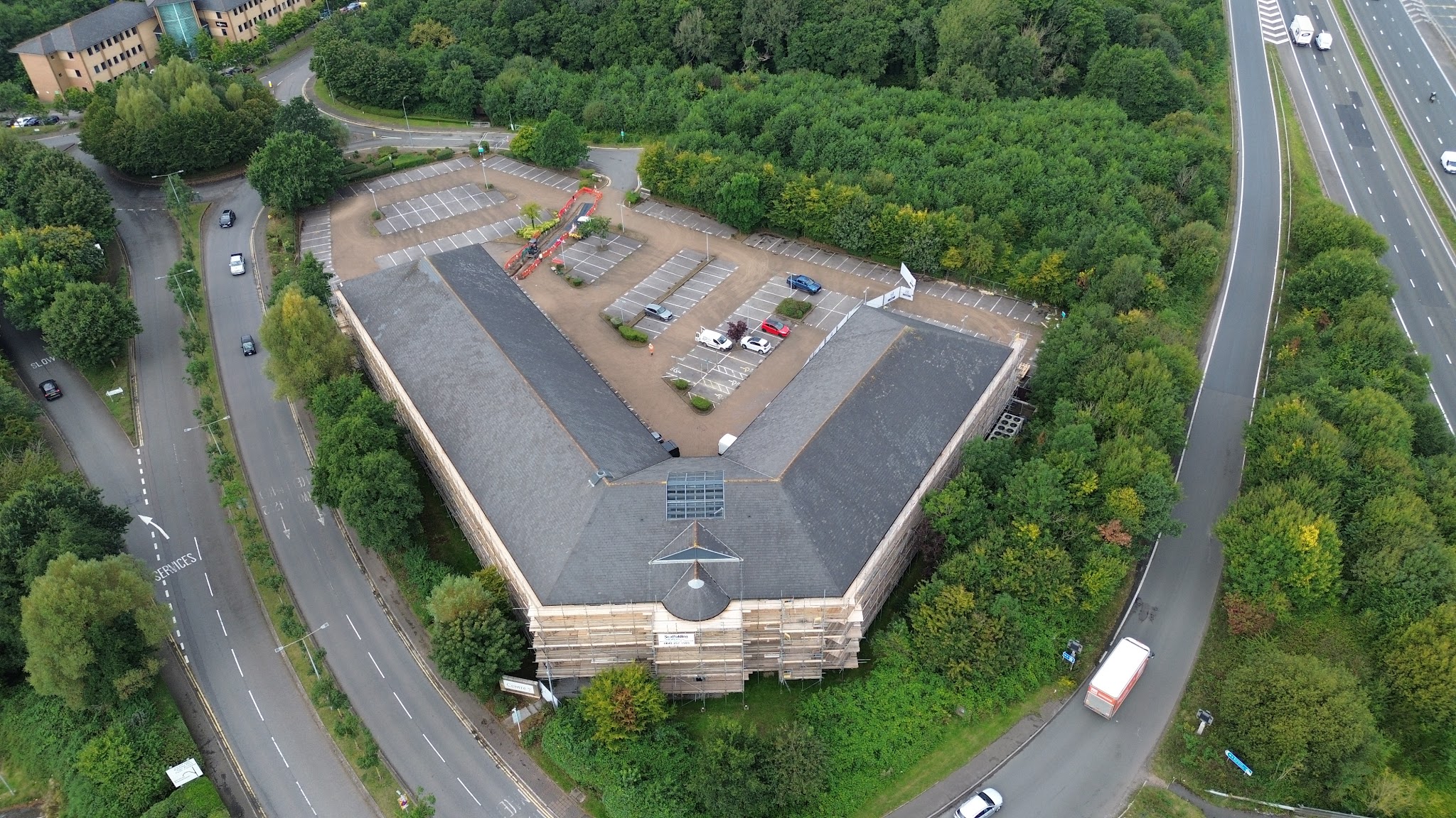 Aerial view of a large office building wrapped in scaffold beside a dual carriageway roundabout