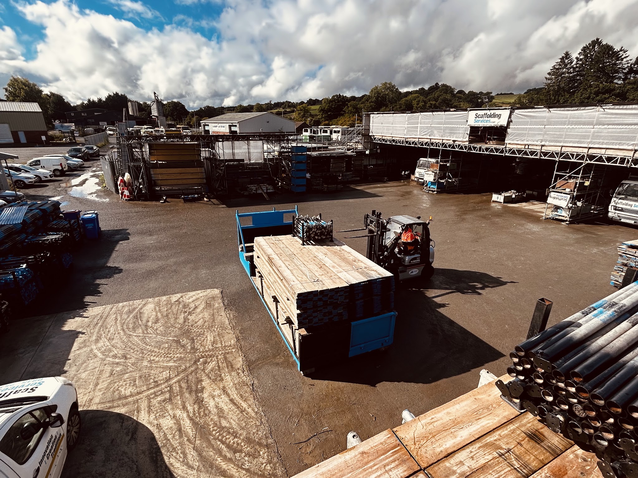 Our yard at Three Cocks: a forklift loading scaffold boards onto a flatbed beside racks of tube and Scaffolding Services vans