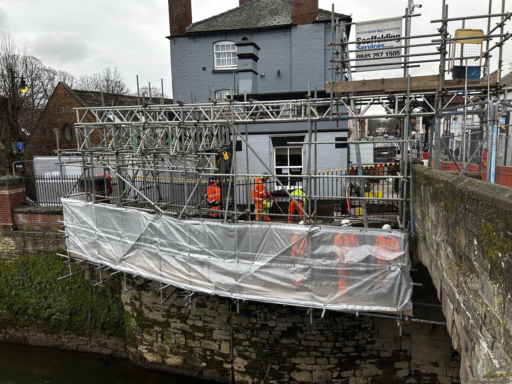 Scaffold with a sheeted lift along a river wall in front of a white building, scaffolders in orange