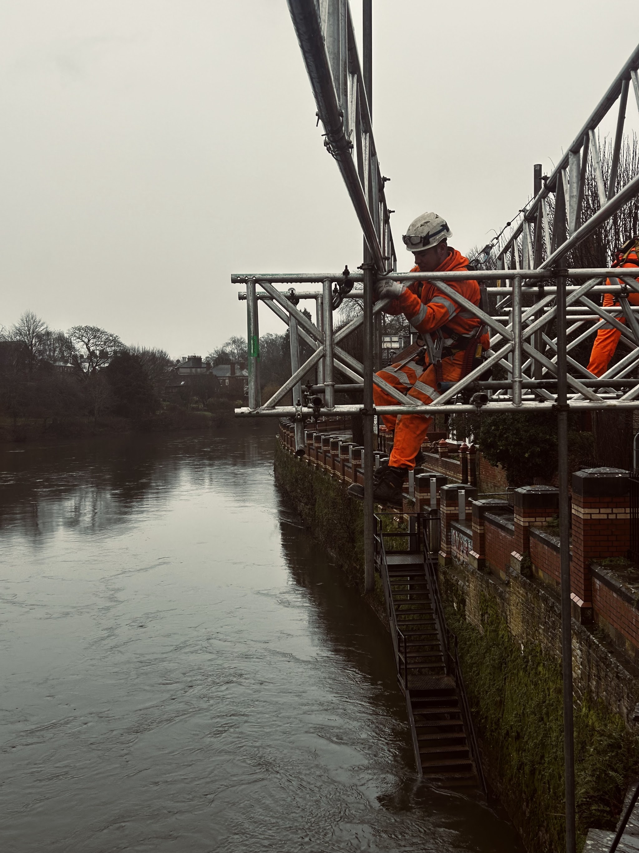 A scaffolder in orange climbing a scaffold built out over a river, beams cantilevered from the wall