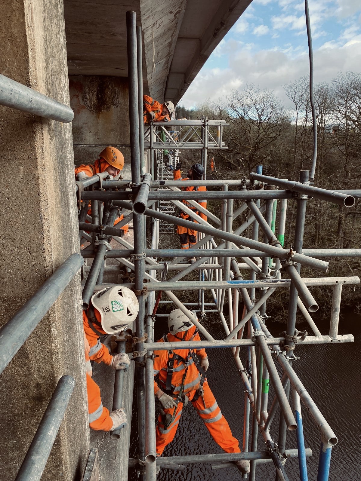 Scaffolders in orange hi-vis and white helmets building tube and fitting under a bridge deck