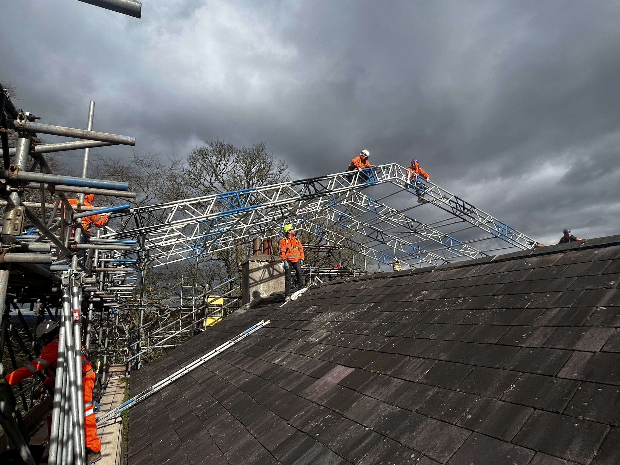 A temporary roof truss going up over a slate roof, scaffolders on top under a stormy sky
