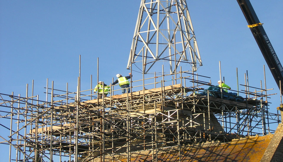 A new church spire being lifted in by crane onto a scaffold platform, workers on the deck