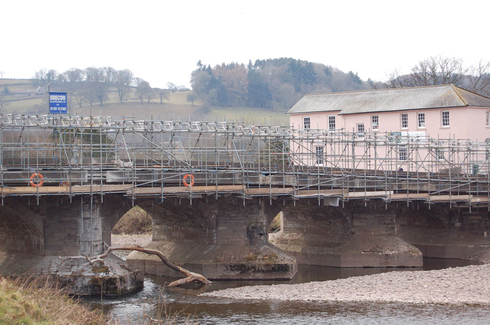 A hanging scaffold under the arches of Llanfaes Bridge over the river, a Brecon Scaffolding banner on the rail