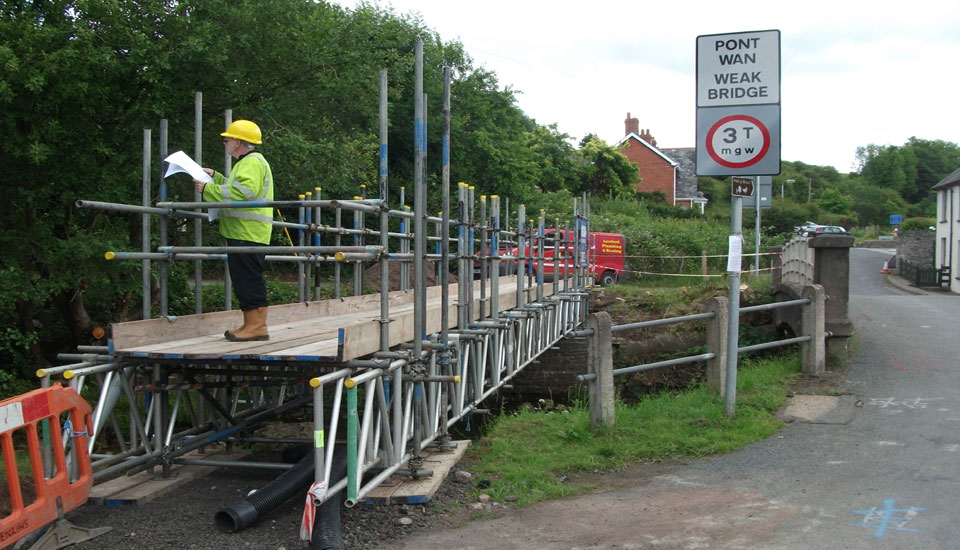A temporary scaffold footbridge with handrails beside a weak bridge sign, a man in hi-vis reading drawings